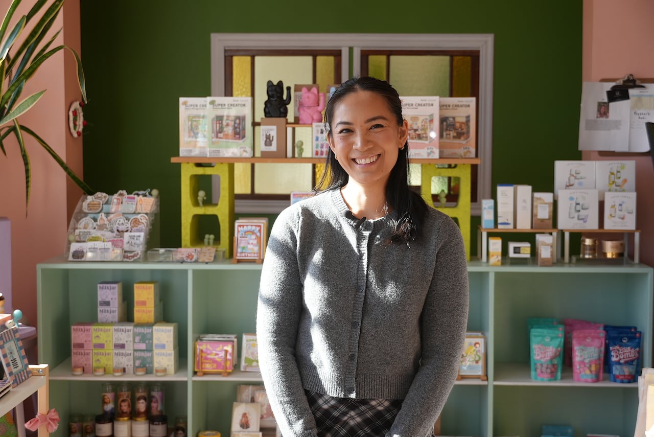 A woman stands smiling inside of her store, with products behind her on shelves. 