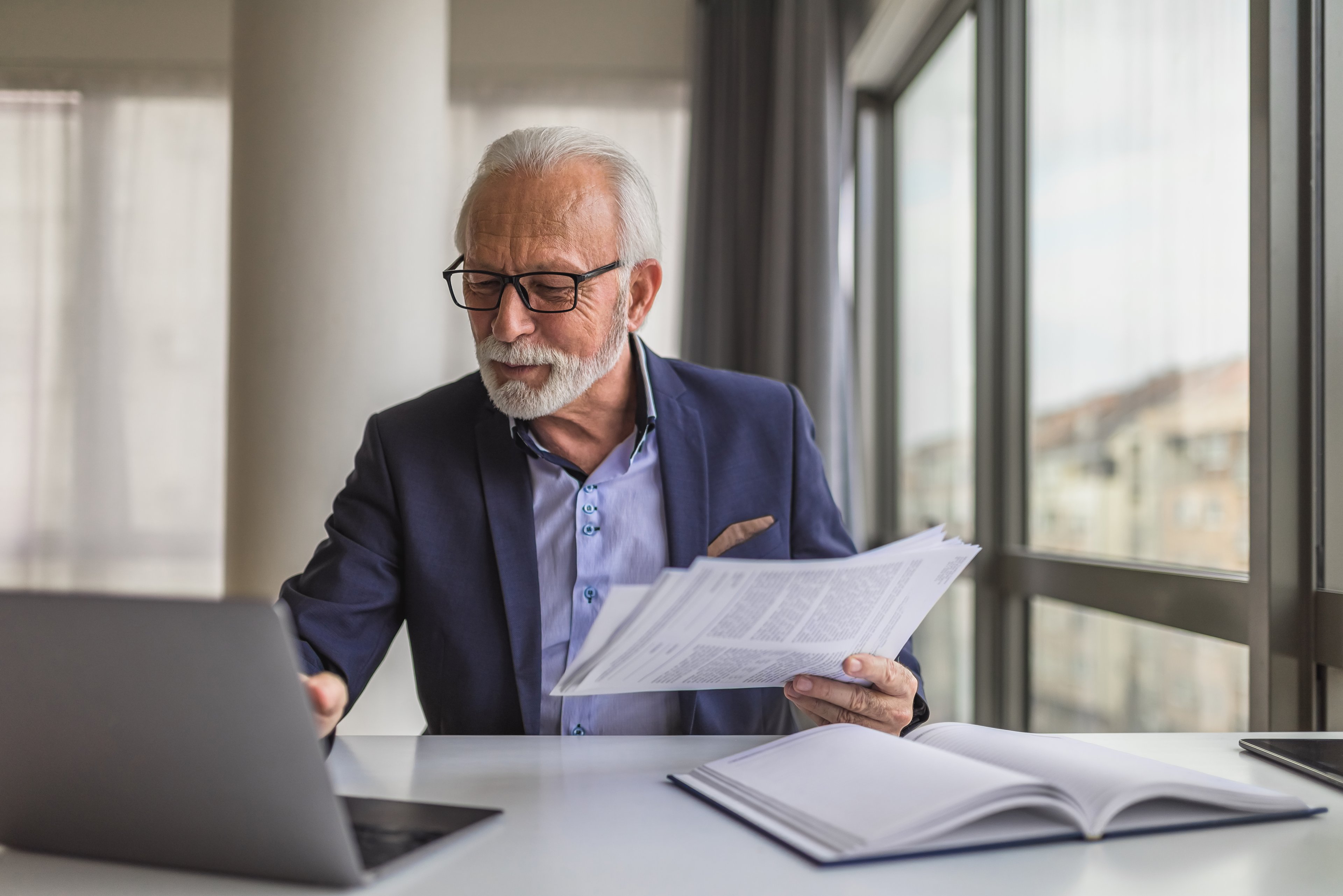 Person in office, holding documents and looking at laptop.