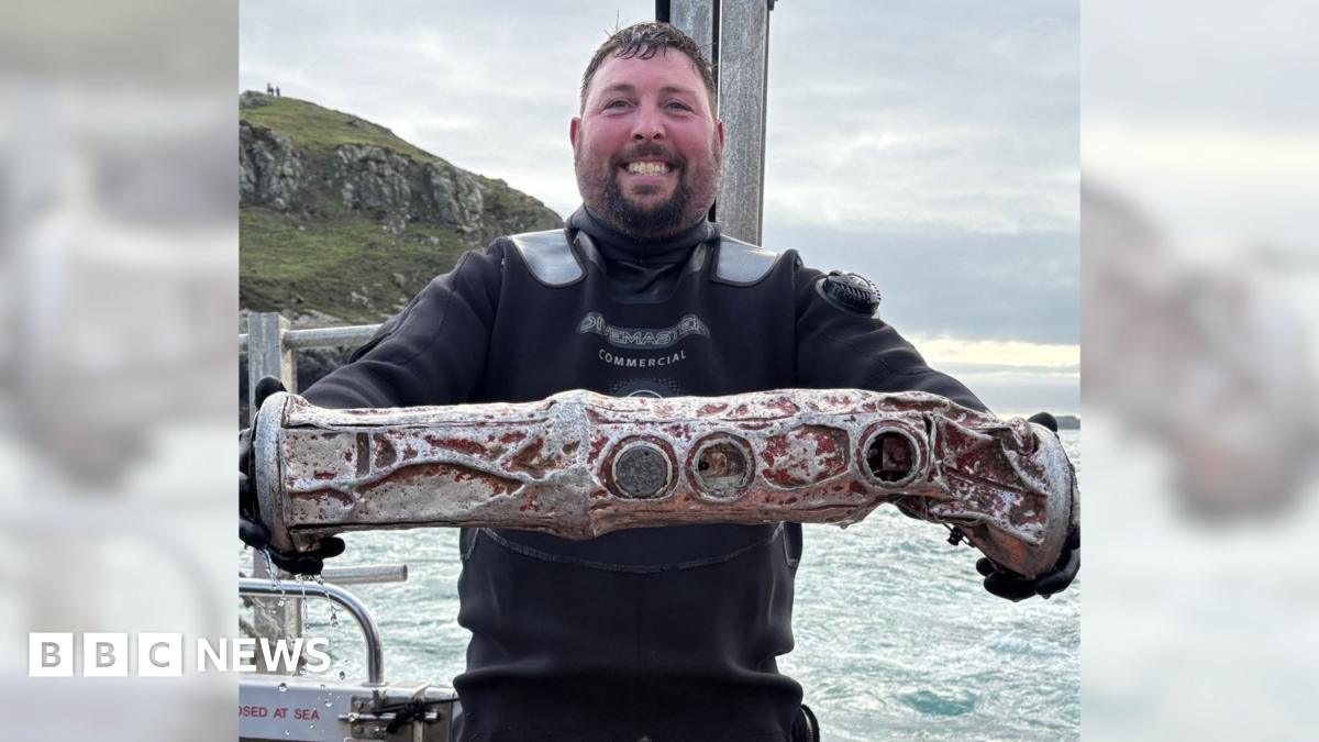 A man in a black wetsuit smiles as he holds a cylindrical metal object believed to be a sonobuoy. He is stood on a boat with, the sea and a portion of land can be seen behind him. He has short brown hair and a brown goatee style beard.