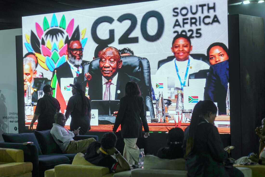 People walk by a large screen TV where South African President Cyril Ramaphosa holds a wooden gavel as he officially closes the G20 leaders' summit, in Johannesburg, South Africa, Sunday, Nov. 23, 2025.