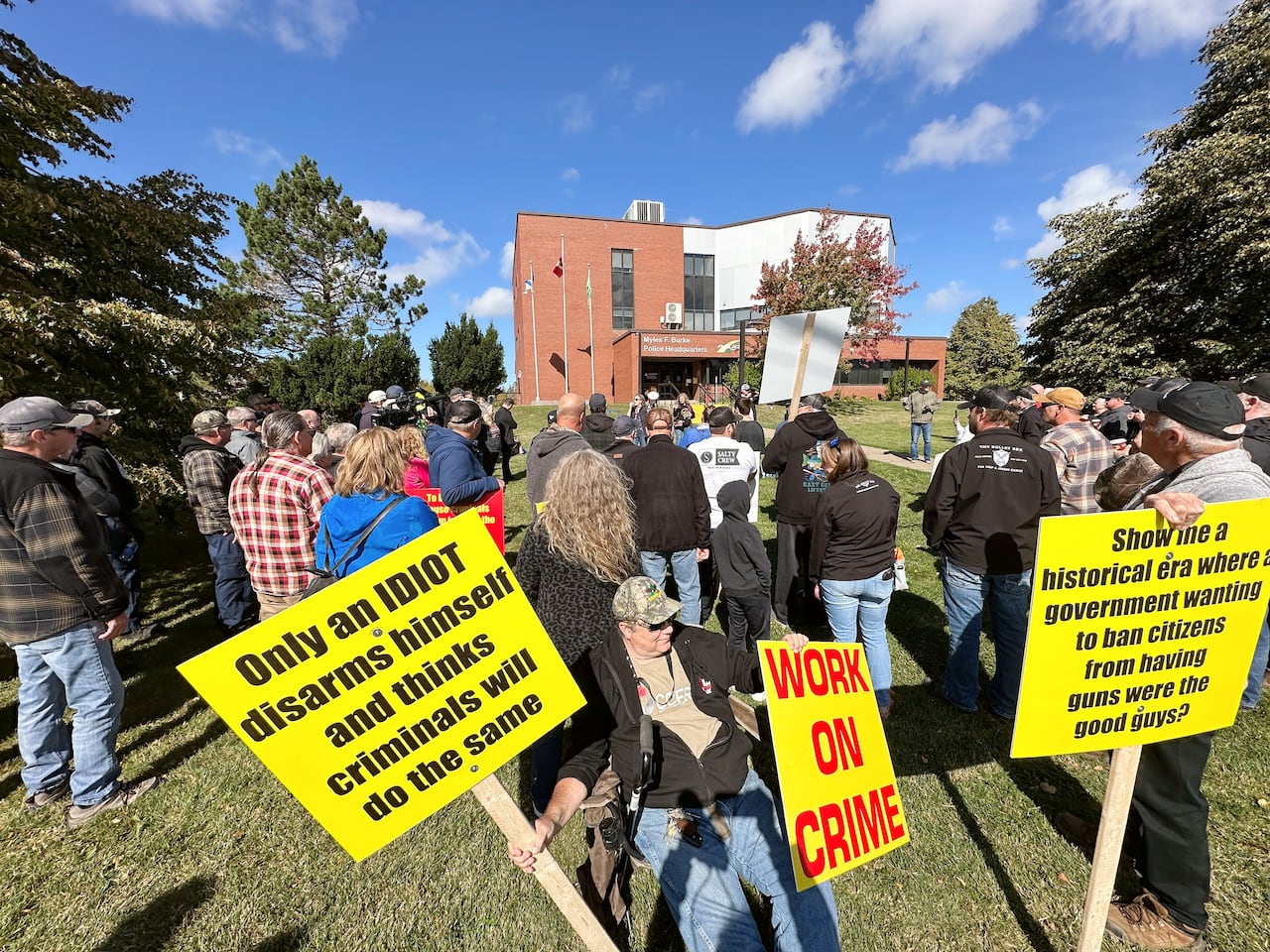 People holding colourful signs stand and sit on green grass in front of a large red brick building with blue sky and white clouds overhead.
