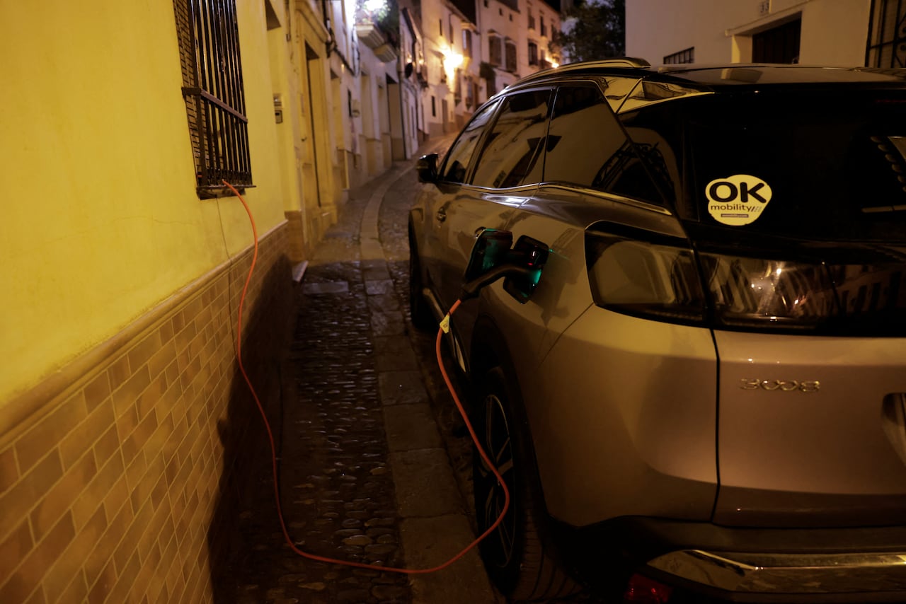 A car is parked on a cobblestone street at night while plugged into an electric charging cable. 