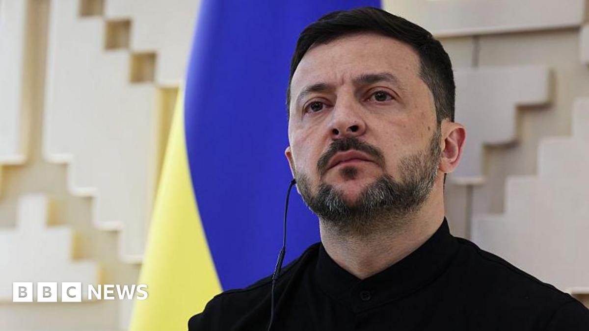 Ukraine's president Volodymyr Zelensky looks out during a press conference. His face is stern. He is wearing a black jacket and shirt. The Ukrainian flag - in blue and yellow - is displayed on the wall behind him.