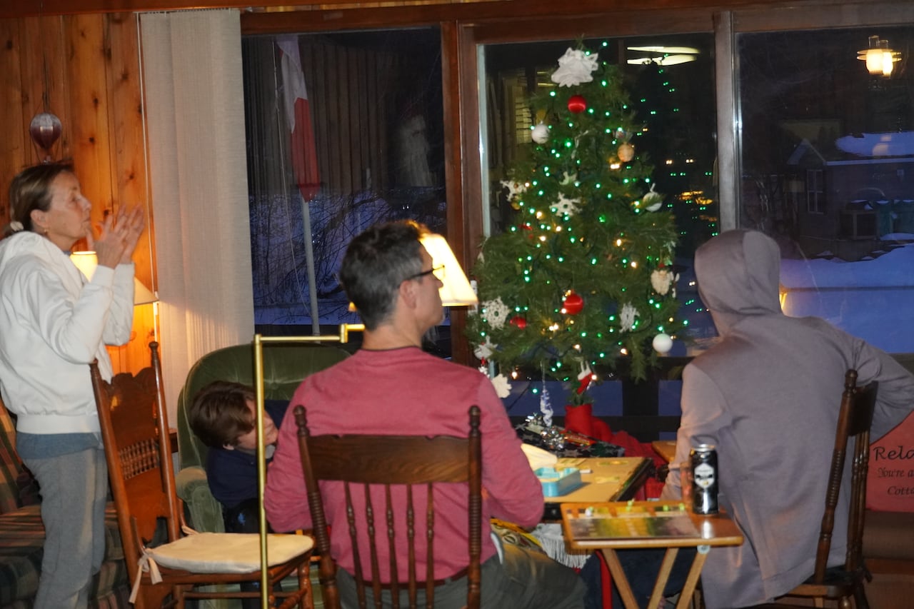 four people sit and stand around a table inside a cottage with wooden panelling on the walls. A Christmas tree is set up. And there is snow outside a window.