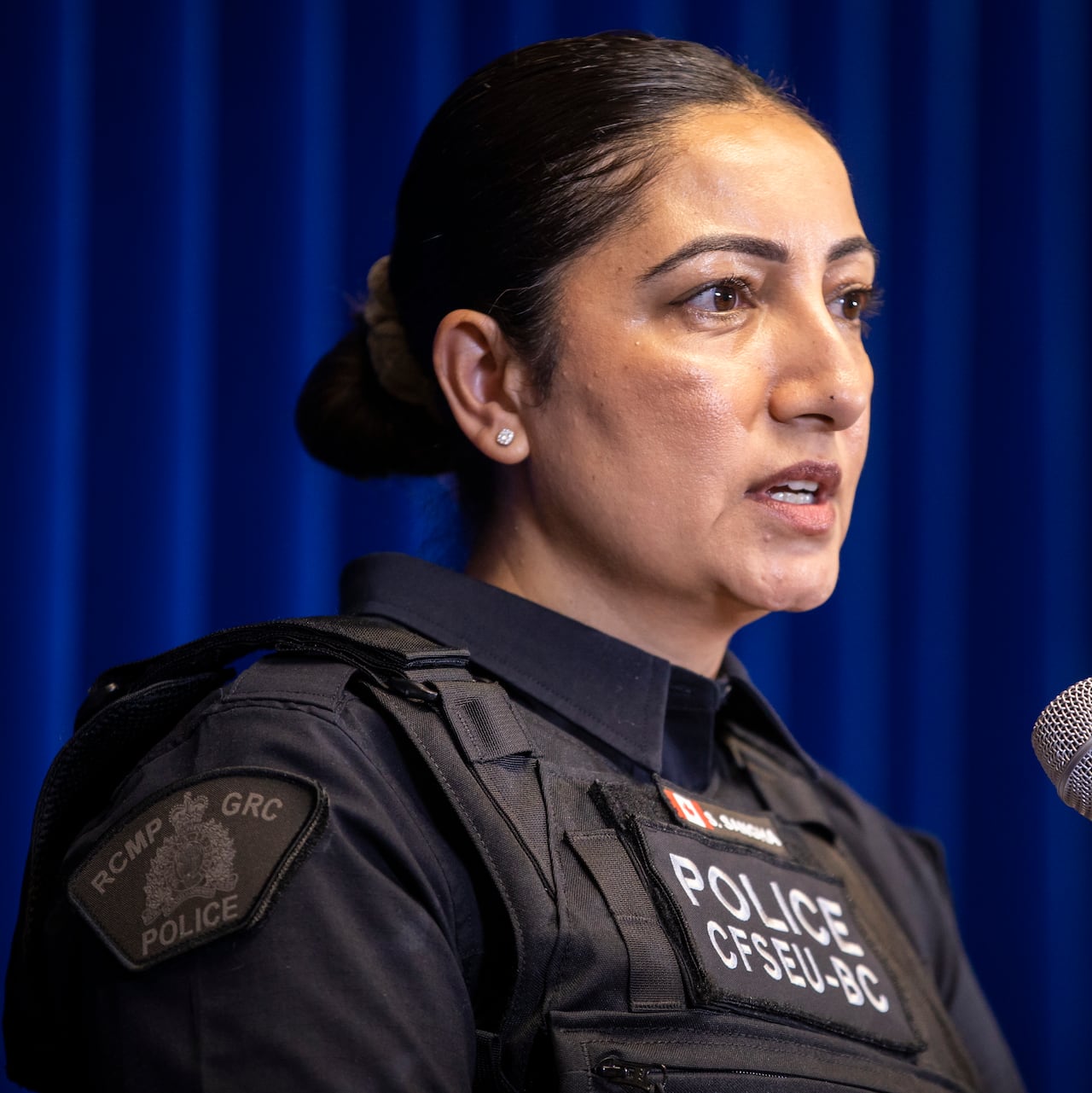 A South Asian woman with her hair in a bun speaks into a mic.