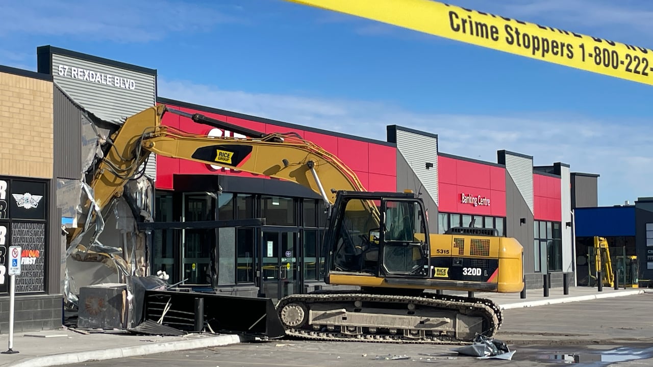 At an empty strip mall parking lot in day time, a yellow excavator is crashed through a CIBC bank, taped off by police tape