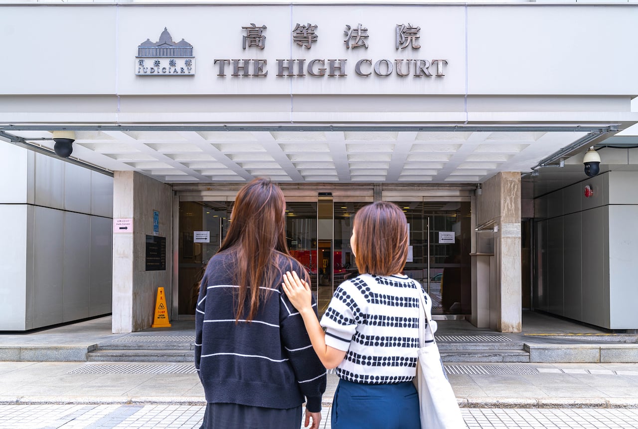 Two woman stand with their backs to the camera, facing a building with a sign that says 'The High Court.'