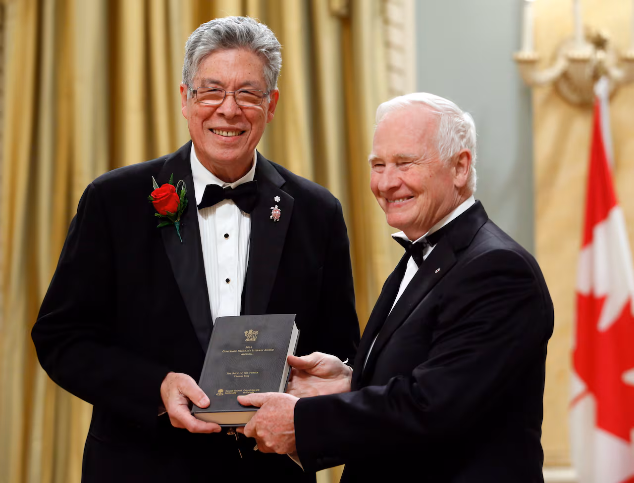 Two smiling, gray-haired men in black tuxedos stand in front of the Canadian flag holding up a hard-cover book. 