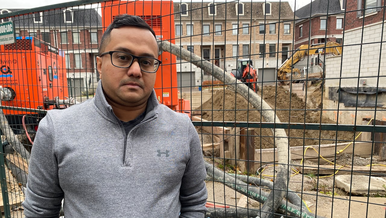 A man stands in front of a fence, with construction equipment visible behind him. 