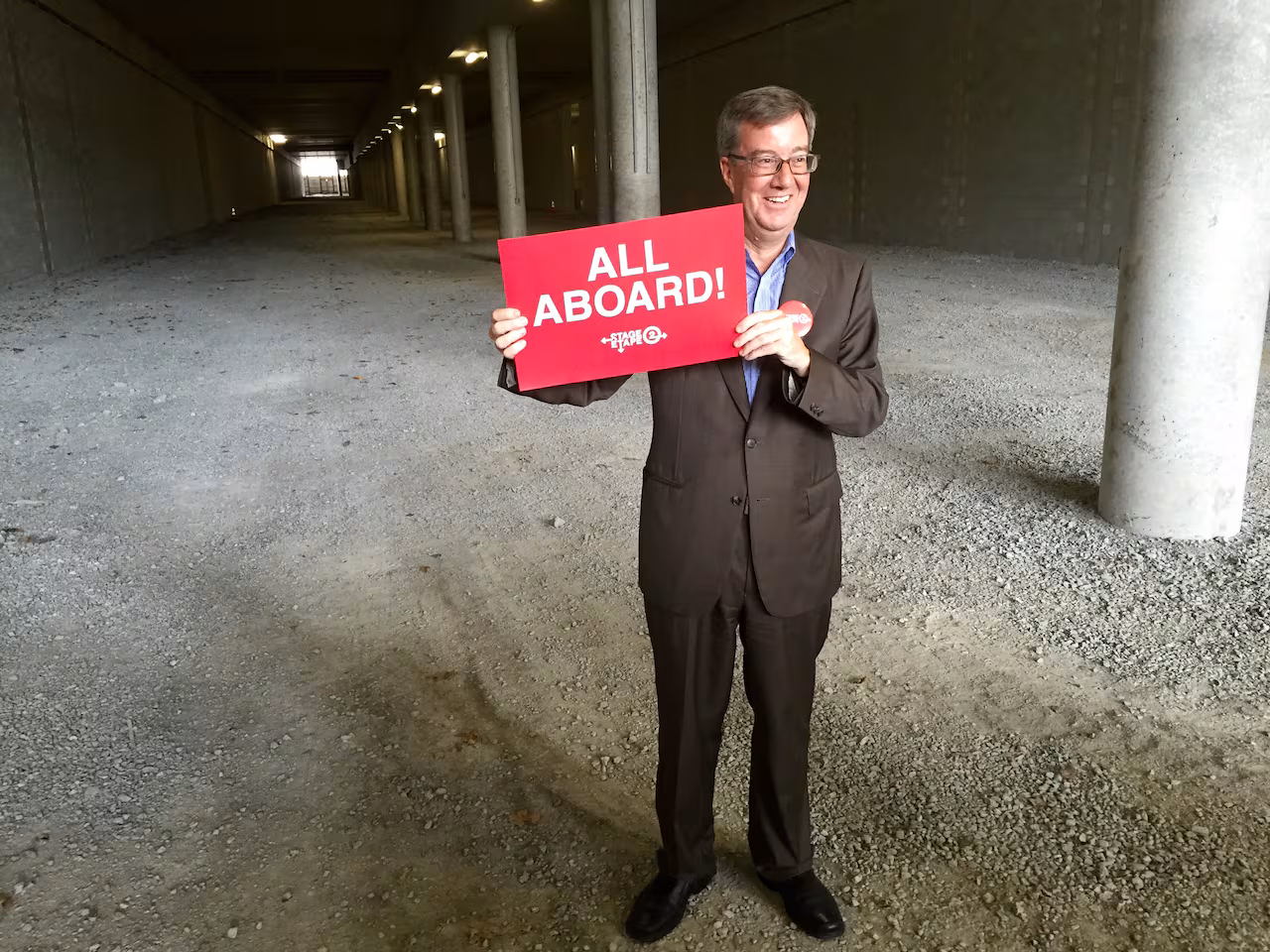 A man in a suit stands in a tunnel holding a sign saying "All Aboard!"