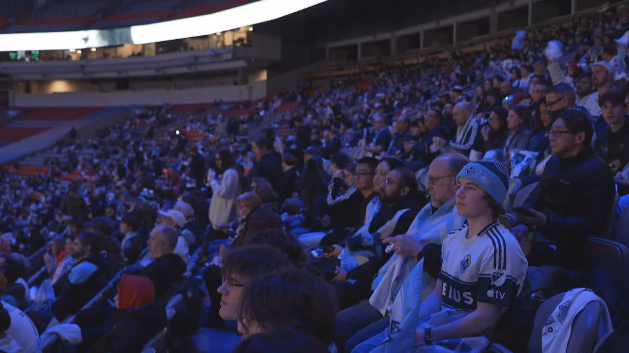  Hundreds of people fill the seats at a stadium wearing jerseys and scarves, watching the match on the stadium screens.