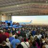This photo shows a scene from a Kamala Harris and Tim Walz rally in Detroit on Aug. 7. Many people are crowded together in an aircraft hangar, facing a stage where Harris speaks at a lectern. The hangar is open to the outdoors, and outside is Air Force Two and a blue sky.