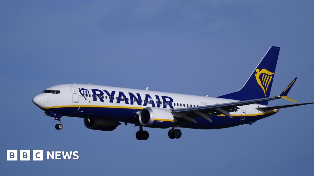 A Ryanair flight seen flying against a blue sky with the gold harp-style branding as its livery on the back of the plane and Ryanair written on its side
