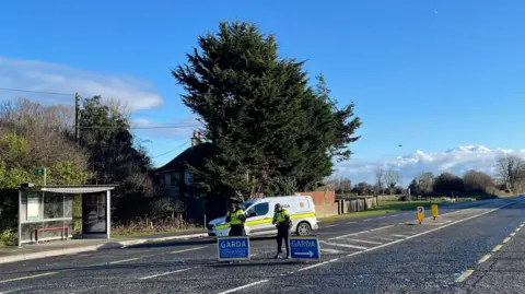 PA Media Gardaí at the scene where a road is closed near where a bus, lorry and car collided in Gormanston, Co Meath. There are two officers standing behind Garda signage. The signs are blue.