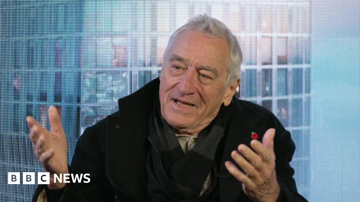 Robert De Niro, sitting in front of a blue background, wears a black coat and a dark scarf. He has short grey hair and is gesturing with both hands out in front of him.