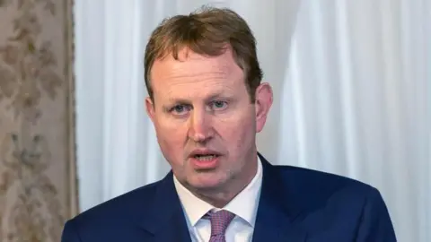 PA Media A man with short brown hair and blue eyes, wearing a blue navy suit jacket, white shirt and pink tie.