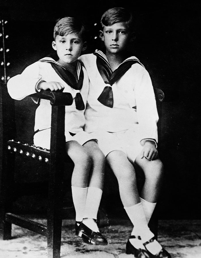 Young King Juan Carlos and his brother sitting side by side wearing sailor outfits in a vintage black and white photo.