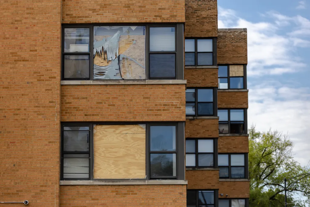 Broken and boarded up windows on a tan-brick apartment building with multiple floors.