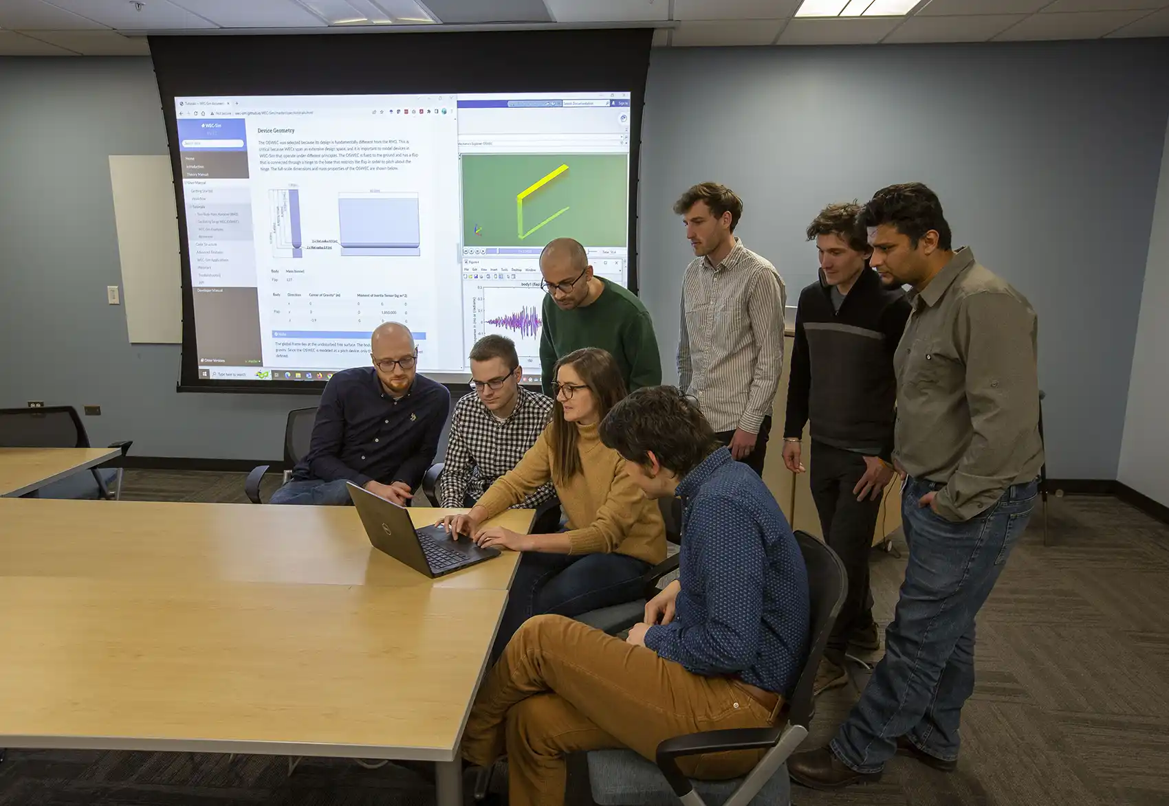 Eight people crowded around a laptop on a table while a screen behind them displays graphic illustrations of a marine energy device.