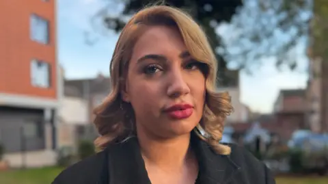 Jo Black/BBC Amira Ostalski is in the middle of the image looking towards the camera. She has shoulder length light brown hair and is wearing a black coat. 