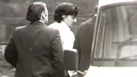 BBC A black and white still from TV footage showing Peter Sullivan, who has thick black hair and is wearing a white shirt, being led to a police van in handcuffs.