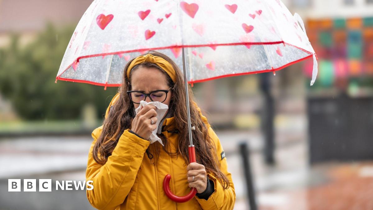 A woman blowing her nose and sneezing into a handkerchief during a rainy autumn day. She is wearing a yellow rain coat and a red umbrella