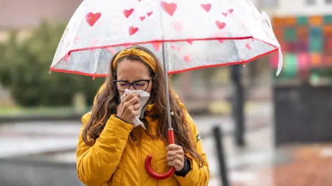 Getty Images A woman blowing her nose and sneezing into a handkerchief during a rainy autumn day. She is wearing a yellow rain coat and a red umbrella 