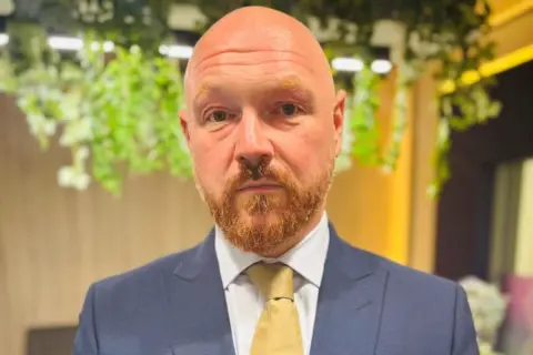 Graham Skinner, a man with a beard, wearing a blue suit, white shirt and yellow/gold tie looking at camera.
