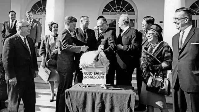 JFK and staffers surrounding a turkey on a pedestal with a sign that signs "good eating, Mr President!"