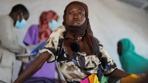 Reuters Halima Mohammed Adam, a Sudanese cholera patient with a brown shawl over her head and wearing a multi-coloured dress, looks on as she sits on a bed at a United Nations-run makeshift clinic in Tawila in Darfur, Sudan, on 5 August 2025