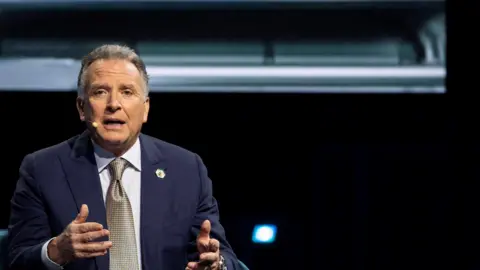 Reuters A middle-aged man in a blue jacket, white shirt and gold tie gesticulates in a room.  