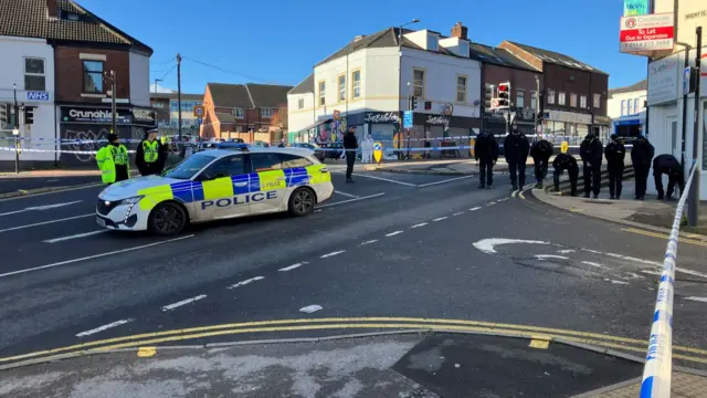 A police car is parked inside a cordoned off area of a Sheffield street. Police officers stand beside it and examine the ground.