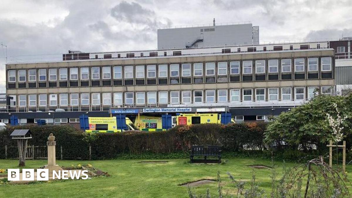 A long, wide building with numerous windows stands in the centre, with ambulances outside an entrance, a sign reads 'Darlington Memorial Hospital'. A park sits in front.