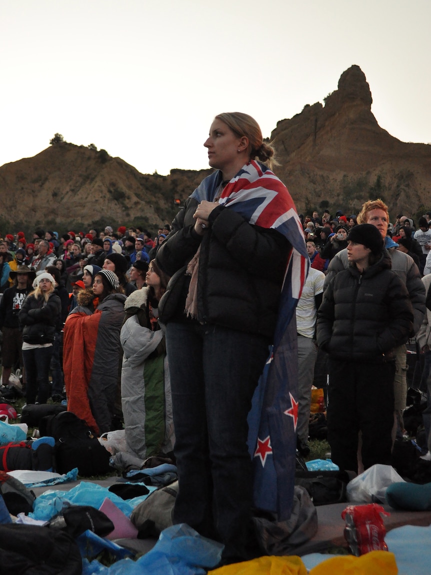 A woman stands in a crowd of people outside by some cliffs, draped in a New Zealand flag