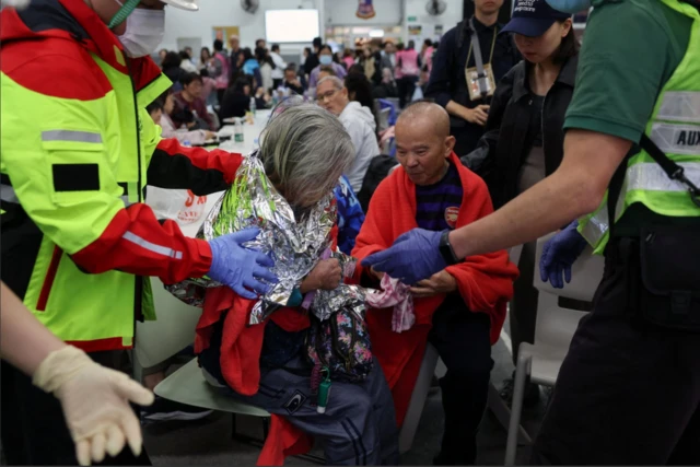 A man and a woman sit down on temporary bedding in a shelter as they are attended to by emergency services. A large group of people can be seen sitting around them
