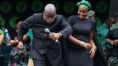 Gallo Images via Getty Images  Jacob Zuma with Duduzile Zuma-Sambudla  dancing outside court. Jacob Zuma is caught in mid-move holding a microphone, looking down and puffing his cheeks. Zuma-Sambudla is smiling and glancing down to her father's feet.
