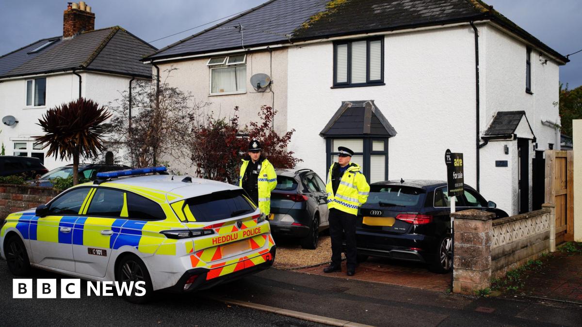 A police car, partially parked on the pavement, outside a white-painted, semi-detached house with black trim on the windows. There is a grey sports car parked on the gravel drive in front of the house, where two police officers in hi-vis yellow jackets stand.