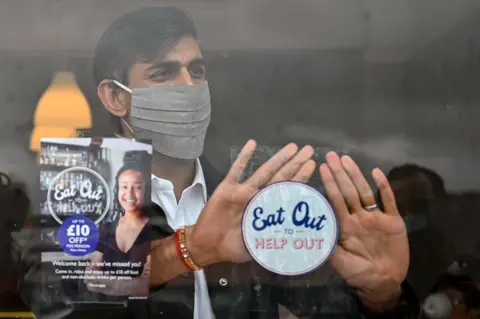 PA Media Photograph of Rishi Sunak wearing a mask in the summer of 2020. He is behind a window of a restaurant and attaching a sticker which promotes the Treasury's Eat Out to Help Out scheme.