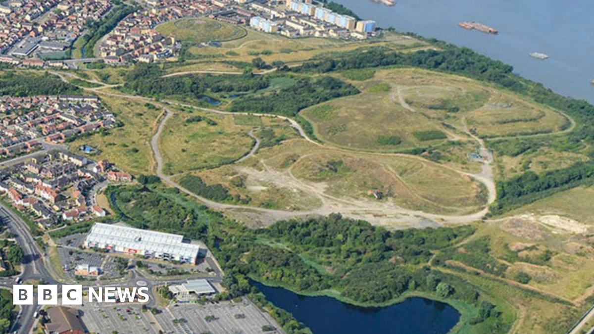 The river Thames can be seen on the right from an aerial shot - with green fields and trees, on the outskirts of which are low and high-rise housing and buildings with a lake in the foreground.