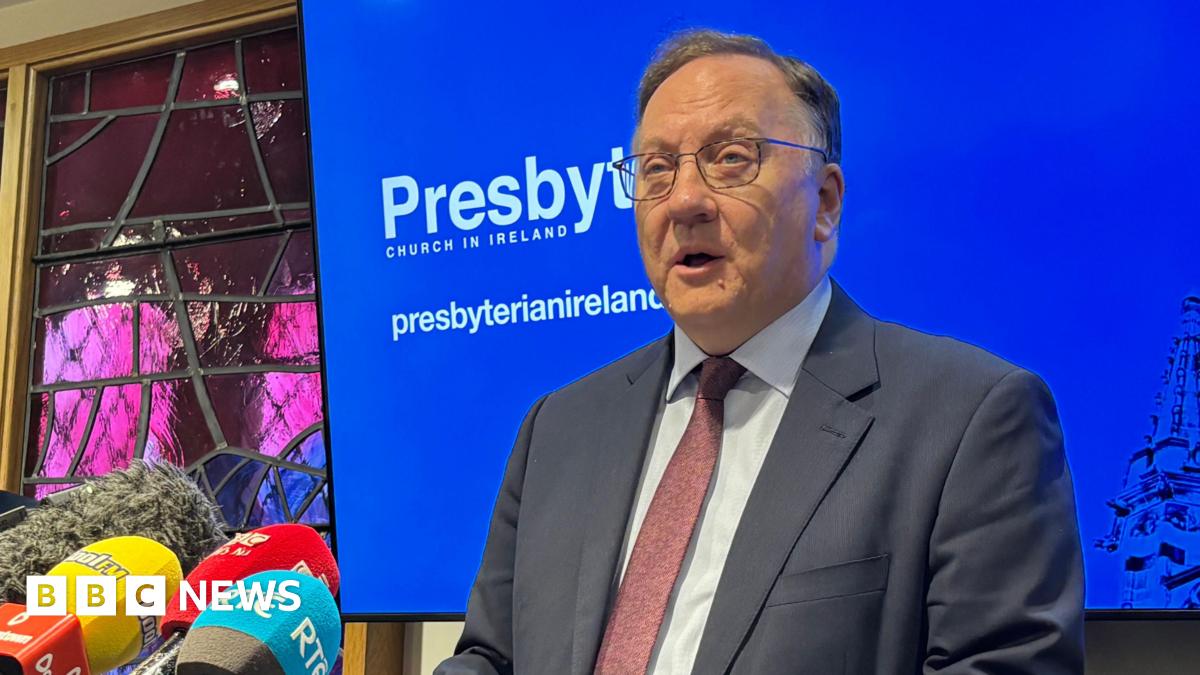 Rev Trevor Gribben standing at a podium with a row of microphones in front of him. There is a stained glass wall behind him, as well as a blue TV screen which says Presbyterian Church in Ireland. He has glasses and is wearing a suit