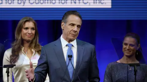 REUTERS/Brendan McDermid Andrew Cuomo stands at a podium with a blue background and two woman and a man behind him.. He is in a grey-blue suit with a blue tie and is gesturing and looking downcast.