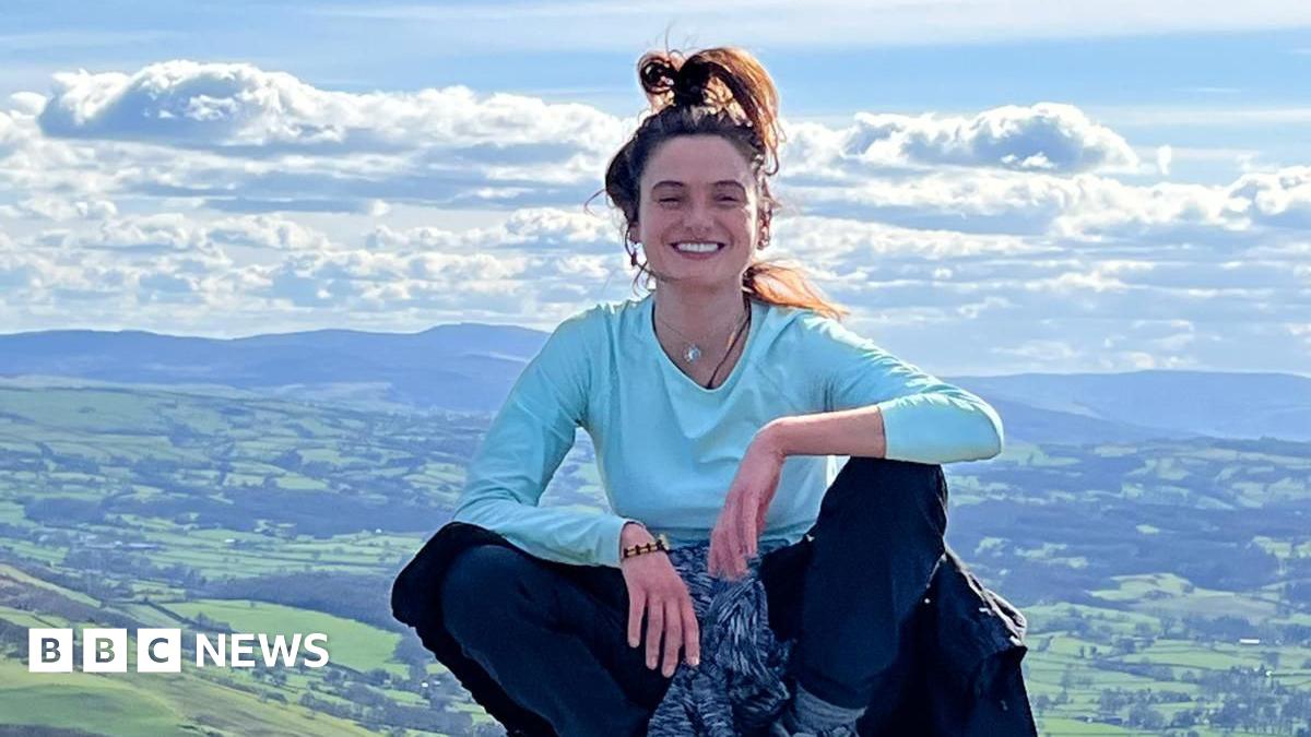 Eleanor Thompson is pictured sitting on a rock, with the landscape below, wearing a blue top and jeans, with a jumper tied round her waist. Her hair is tied in a high ponytail and she is smiling.