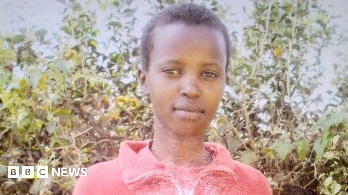 A picture of Agnes Manjiru standing in front of a bush with green leaves. She is wearing a pink cardigan-style top and has short cropped black hair