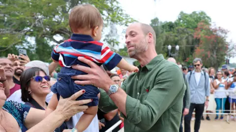 PA Media William wearing a khaki shirt with a poppy pin attached to the pocket and he holds a baby boy with both hands. The boy has his back to the camera and is wearing a horizontally striped tshirt in navy, red, turquoise and white, and navy shorts. The boy is being handed to William by a woman who is only in shot by her hands. They are among the crowds of people behind barriers waiting to catch a glimpse of the prince
