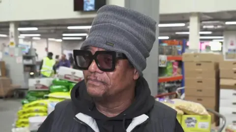 Reggae singer Maxi Priest standing in front of a grocery shop. He is wearing a grey wooly hat and large black sunglasses, as well as a hoodie and gilet.