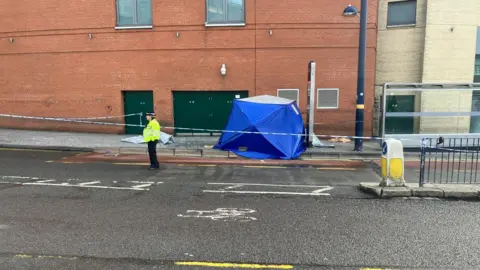 A blue police tent on a pavement surrounded by blue and white police tape. A police officer stands guard wearing a black hat and yellow hi-vis jacket
