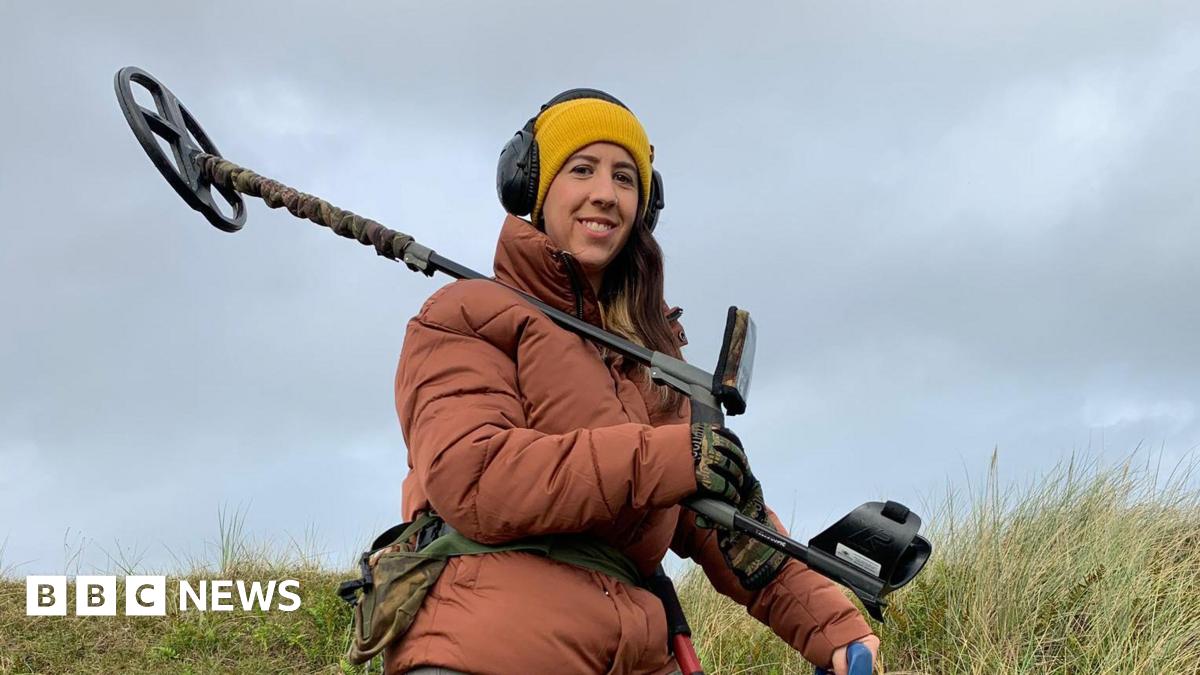 Woman in orange coat, yellow hat and headphones holds a metal detector over her shoulder. She smiles at the camera, army print gloves on and matching bag around her waist. She stands on grass, the sky is overcast.