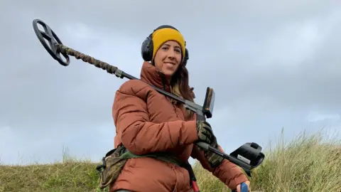 Handout/BBC Woman in orange coat, yellow hat and headphones holds a metal detector over her shoulder. She smiles at the camera, army print gloves on and matching bag around her waist. She stands on grass, the sky is overcast.