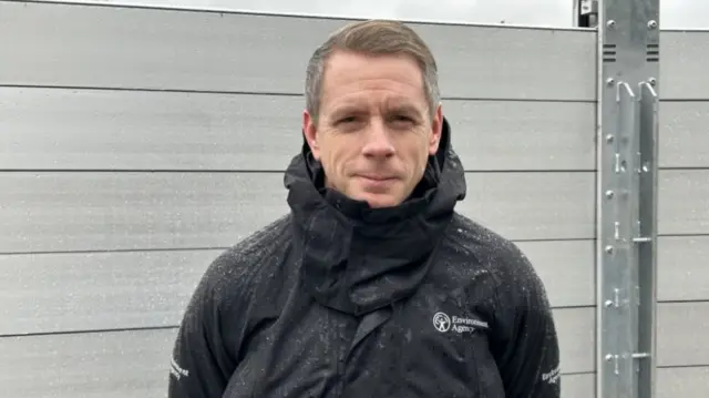 A man with brown hair and a black waterproof jacket standing in front of a metal wall