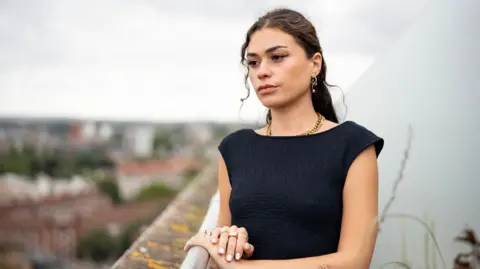 BBC/SpunGold TV Elle, a young woman with long, dark hair, wearing a black sleeveless dress, with golden earrings and a golden necklace, looking pensive as she leans against the railing of a rooftop balcony and looks out at the city below on an overcast day.